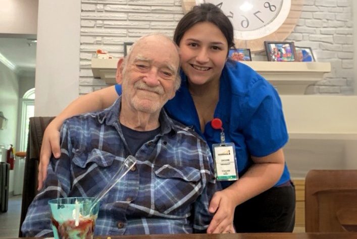 Elderly man in a plaid shirt smiling with a young female caregiver in a blue uniform at an assisted living facility.